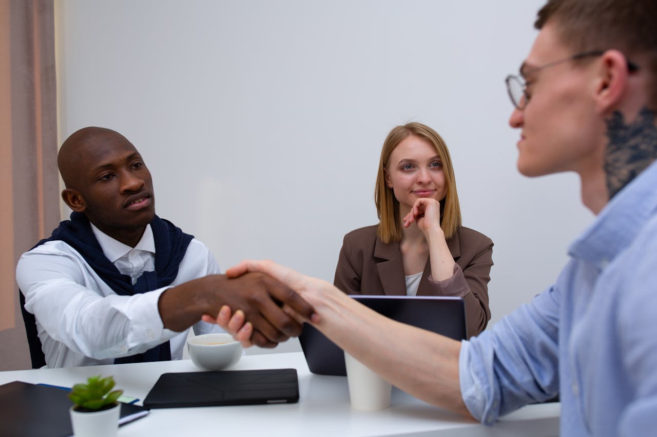 Business professionals in a meeting shaking hands across a white table in a modern office.